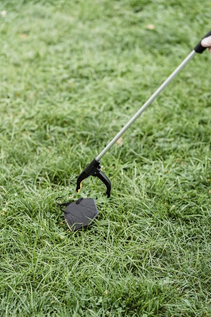The image shows a metal detector probe with a black circular coil at its tip, being used to scan lush green grass outdoors. The grass appears dense and well-maintained, with no distinct flowers or flo