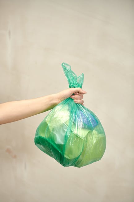 A person’s hand holds a green and blue biodegradable floral waste bag, designed for responsible disposal of floral materials, against a neutral beige wall. The bag appears full and crumpled, indicat