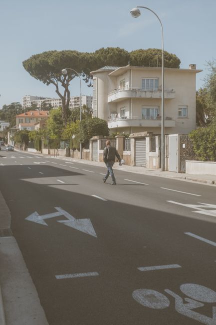 The image shows a city street scene with a person crossing the road near a large, modern, cream-colored residential building that has multiple balconies. Tall, umbrella-shaped pine trees line the stre