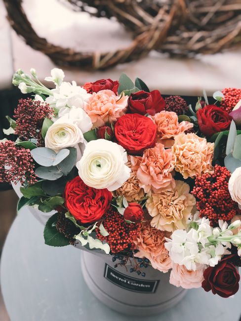 A vibrant floral bouquet featuring red roses, white lisianthus, peach carnations, and small dark red berries, arranged with green eucalyptus leaves in a round white hatbox. The bouquet is displayed on