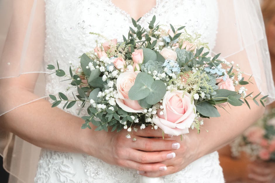 A person in a white wedding dress holding a floral bouquet arrangement featuring soft pink roses, white baby's breath, and eucalyptus leaves. The bouquet has a lush, rounded shape, with the flowers ar