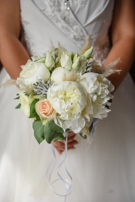 A bridal bouquet featuring a harmonious arrangement of white and pale pink roses, peonies, and lisianthus, accented with soft, grey-green foliage and delicate feather-like textures. The bouquet is hel