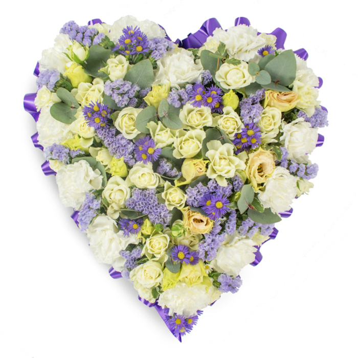A lovingly assembled heart-shaped floral tribute photographed against a pristine white background, where every element is chosen to convey calm and remembrance. The core of the heart is a lush tapestry of cream and pale yellow roses, their layered petals offering a soft, luxurious texture that reads almost like silk when lit by soft studio light. Intermixed are classic white carnations with their frilled edges, placed to create gentle depth and an enveloping, comforting surface. Windings of lavender statice and clusters of vivid purple aster-type flowers, each with a tiny yellow eye, breathe pockets of cool color through the pale base and provide delicate, natural contrast. Throughout the piece, rounded eucalyptus leaves in blue-green tones are positioned to punctuate the design with sculptural clarity and to relieve the floral density with their broader, matte forms. The heart is carefully edged with a continuous, rich purple satin ribbon, folded into neat loops that define the silhouette and lend a formal, respectful finish against the white backdrop. The overall effect is serene and consoling: a bouquet that balances classic sympathy florals with subtle, contemporary accents, suitable for services, wakes, or graveside placement in Somers Town and surrounding streets like Ossulston and Coburg Crescent. The arrangement communicates steadiness and care-soft aromas implied by fresh foliage, tactile petal layers, and a composed, heartfelt statement of remembrance meant to comfort mourners and honour the memory of a loved one.