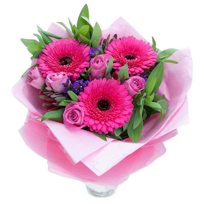 I see this bouquet as a lively little celebration of pinks and purples captured against a bright white surface: three vivid fuchsia Gerbera daisies open fully at the centre, their many petals creating a bold, joyful focal point with deep dark discs that anchor the colour. Nestled close are several soft lavender-pink or dusty-rose roses, petals tightly formed as if pausing between a whisper and a song-fresh, tender, and inviting touch. Interspersed are delicate sprays of purple-blue statice (limonium), whose tiny clustered flowers provide both texture and a subtle cool note that complements the warmer pinks. Surrounding foliage, likely salal or eucalyptus, lends a lush green backdrop; the leaves appear broad and slightly waxy, catching the light in places and adding a crisp, botanical contrast to the velvety petals. The bouquet is carefully wrapped: an inner cushion of pale pink matte tissue paper supports the blooms while an outer layer of translucent, slightly crinkled fuchsia-pink cellophane adds sheen and keeps the arrangement cohesive-edges folded with an experienced hand. Visible at the bottom is the clear glass vase rim, a reminder that these stems are ready for water and will open further on the windowsill. Imagine the scent: a light rose perfume tempered by the clean green notes of gerbera and foliage, a fragrance that lifts the spirit. Perfect for brightening a home near King's Cross, a flat on Ossulston Street, or a table at a café close to Pancras Square, this bouquet feels intimate, joyous, and meticulously crafted for moments of appreciation or celebration.