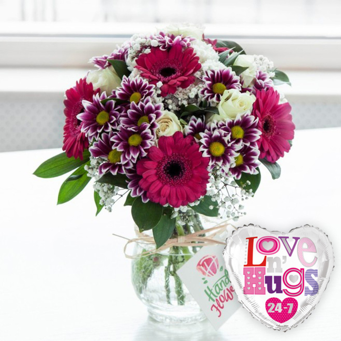 A bright, affectionate composition captures a bouquet and balloon arrangement that feels right at home in the lively yet intimate streets of Somers Town. At the centre is a clear, rounded glass vase placed on a clean, pale surface, its simplicity allowing the flowers to take full focus. Through the glass, straight green stems are visible, immersed in clear water that reflects the light, showing that the bouquet has been freshly prepared. The arrangement forms a generous dome of colour, with large magenta and fuchsia gerbera daisies acting as the stars of the display; their dark, almost black central discs provide striking contrast against the vivid petals radiating outward in fine, even spokes. Clustered among them are smaller chrysanthemums whose petals are deep purple near the centre and fade to crisp white at the tips, while their bright yellow cores offer friendly, cheerful dots of light. Creamy white roses, some still rounded in bud and others beginning to open, are interwoven through the bouquet, their soft, layered petals bringing a classic romantic mood to this otherwise modern colour scheme. Fine clouds of white baby's breath float throughout, lending a delicate, lace-like texture that softens edges and fills tiny gaps, ensuring the arrangement looks full from every angle. Fresh green foliage peeks out around the sides and base, balancing the brighter tones and giving the design a natural, garden-fresh aesthetic that could easily have been inspired by local greenery around Somers Town. Wrapped around the vase neck is a piece of rustic natural twine, tied into a small bow that supports a neat white gift tag decorated with a pink rose motif and friendly green script, suggesting a personal note from a caring florist. To the right of the vase, slightly angled towards the viewer, a heart-shaped foil balloon with a mirror-like silver border and a clean white centre completes the scene. Across the balloon, the phrase Love n' Hugs 24-7 is printed in bold, mosaic-style letters filled with patterns in shades of pink, purple, and blue, accented by tiny heart shapes. The softly out-of-focus background is bright and white, as though the bouquet is standing near a sunlit Somers Town window or studio wall, creating an uplifting, celebratory atmosphere ideal for romantic surprises, birthdays, anniversaries, or simply sending love across the neighbourhood with same-day flower delivery.