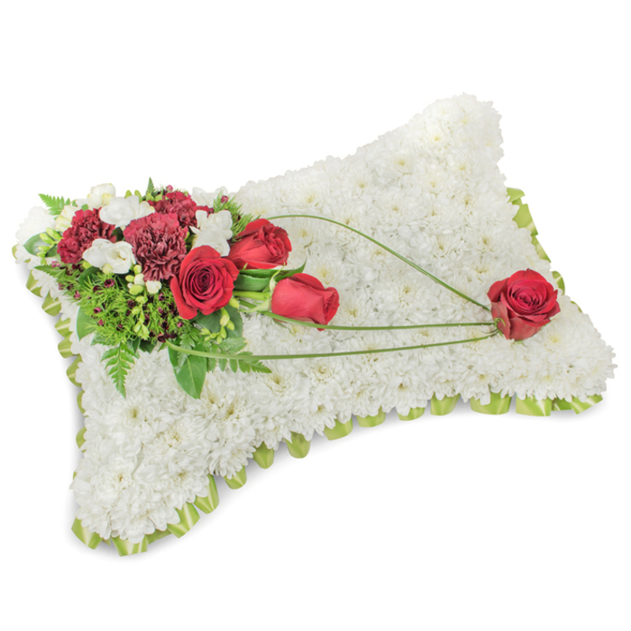 A poignant rectangular funeral cushion formed from a dense carpet of small white chrysanthemums, shown on a neutral white surface so every petal and contour is visible. The chrysanthemum heads are carefully positioned to create an even, velvety texture that suggests comfort and purity; around the softly raised edge a pale green trim - likely ribbon or delicate foliage - peeks through, giving the piece a refined border. In the upper left a concentrated cluster of deep-red flowers provides a striking, heartfelt contrast: several red roses, some tightly furled and others opening to reveal velvety spirals, are paired with darker burgundy carnations whose layered frills add richness. Interspersed small white blossoms, perhaps freesias or spray chrysanthemums, introduce lightness, while a backdrop of mixed greenery, from feathery fern fronds to broader leaves, lifts the composition with natural depth. Three slender, curved lengths of bright green bear-grass or similar foliage trail diagonally from this main spray toward the lower right, articulating a graceful movement that culminates in a solitary, perfectly formed red rose resting at the pillow's corner - an intentional focal point suggesting remembrance and love held close. The bouquet feels calm in scent and presence, suited to services in local venues around Somers Town, from chapel rooms near Euston Road to quiet memorials close to King's Cross, and offers a classic expression of sympathy crafted with respectful attention to detail.